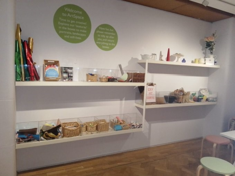 Shelves lined with wicker baskets are full of art materials for children. Two stools and a table are on the right hand side. Signage above the shelves encourage families to get creative and explore the textures in the boxes to make portraits and more
