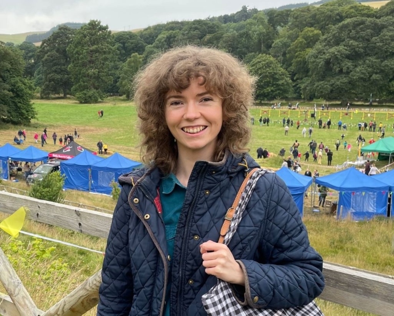 Claire faces the camera smiling. In the background a festival is taking place - groups of people are in a field surrounded by stalls