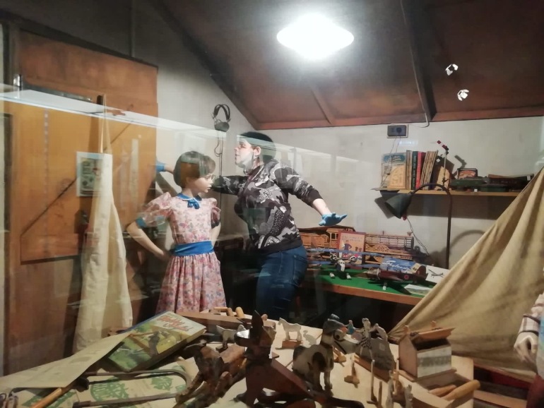 Curator works inside a display case next to a mannequin. The mannequin wears a pink floral dress with blue collar