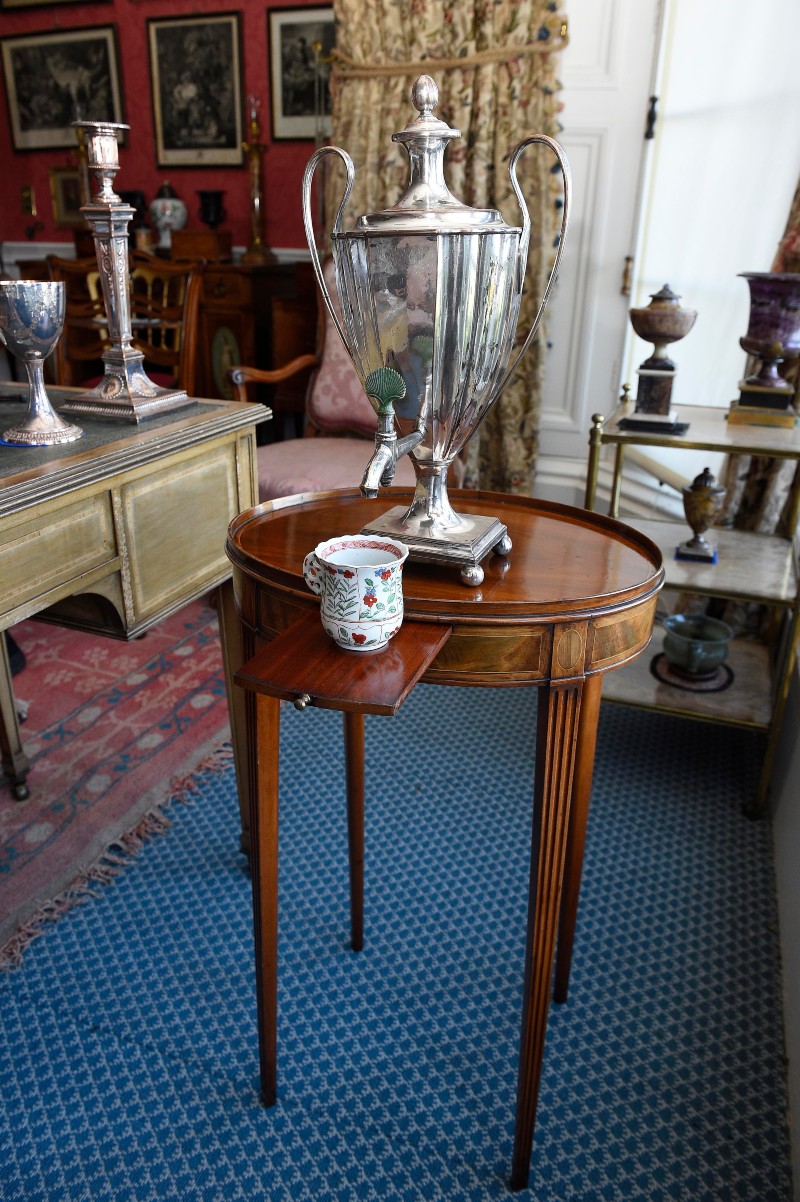 China cup and large polished silver tea/coffee urn sit on top of oval mahogany table 