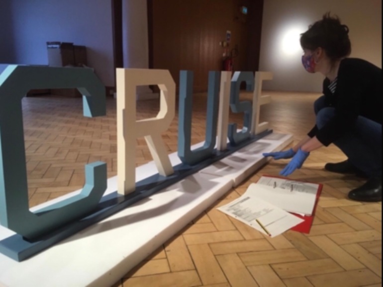 Curator wears blue gloves and kneels beside cut out lettering in blue and white spelling Cruise. 