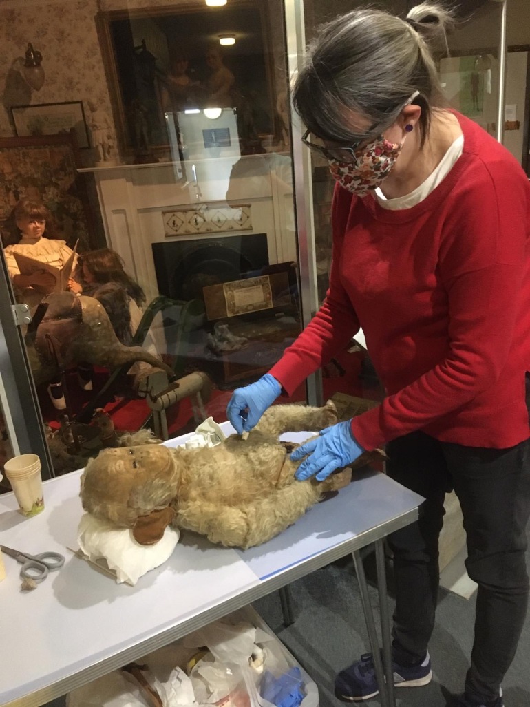 Curator wears red jumper, floral mask and blue gloves whilst cleaning a vintage toy monkey