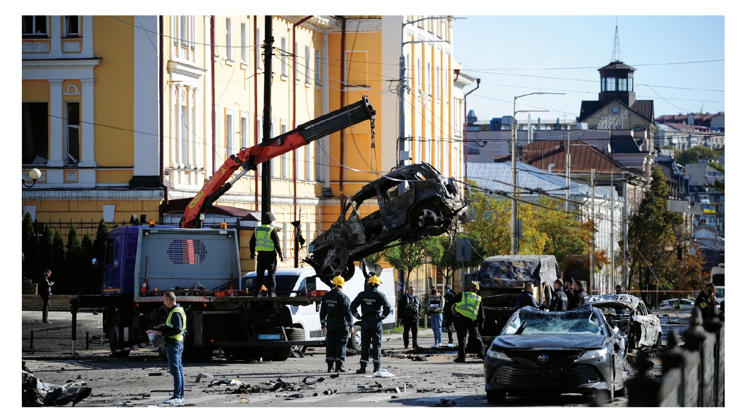 The effects of a missile strike on a street in Kyiv. Damaged cars line the street while a crane uplifts a car and a team of emergency service workers supervise