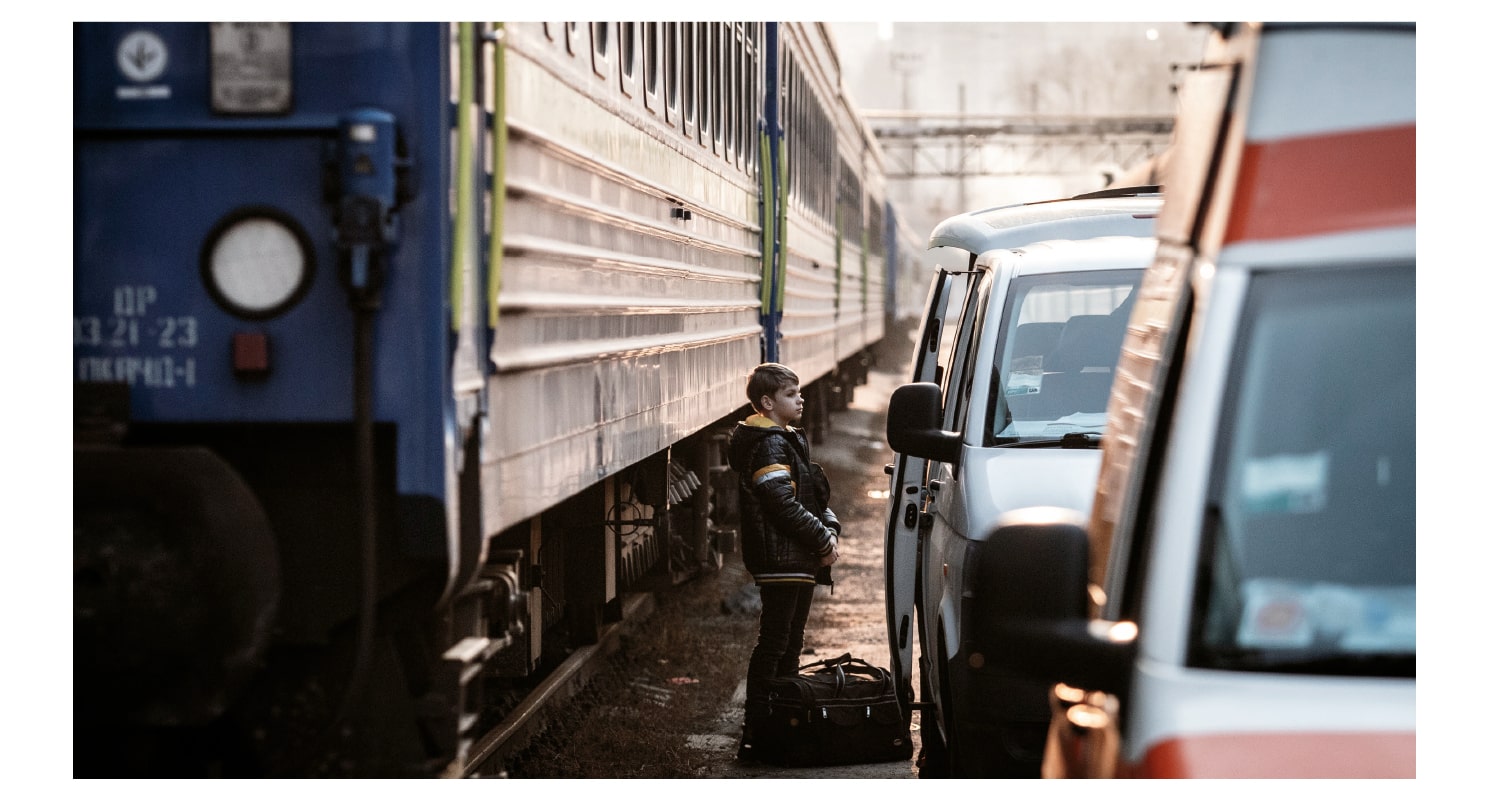 A little boy, with a bag at his feet, stands beside a van and a train and waits for evacuation