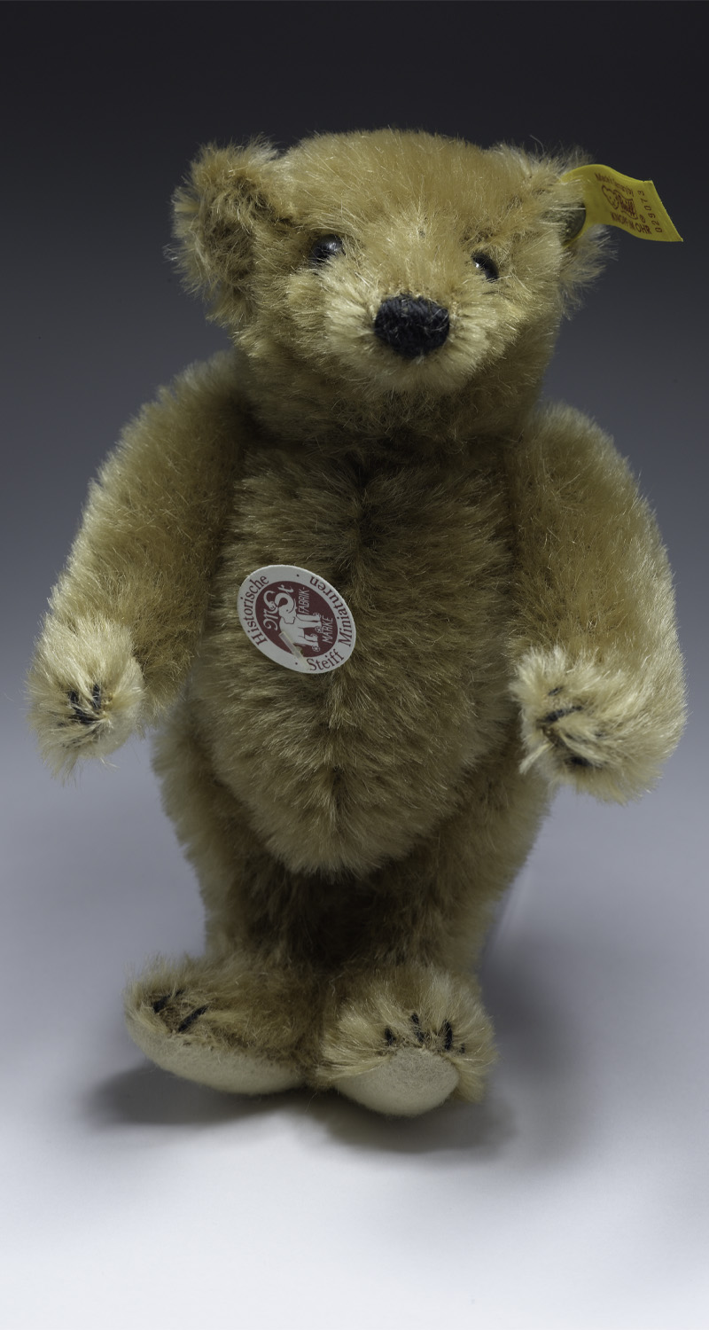 A light brown coloured teddy bear standing up on his back legs. He has a badge with the words "Historische Steiff Miniaturen"