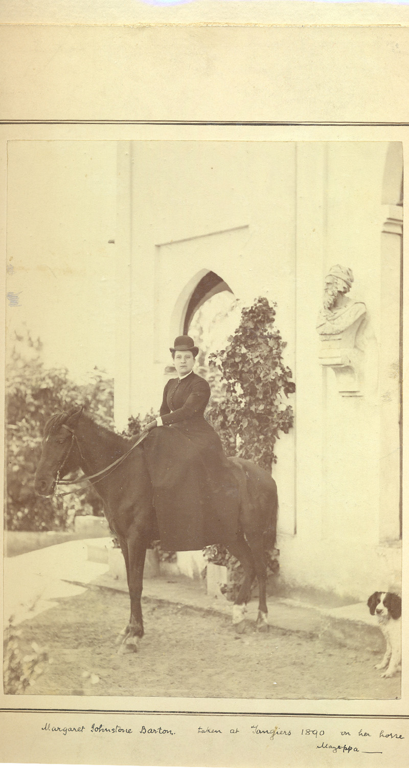 a woman dressed in black riding a horse with a small dog at the horse's feet in front of a large house with pointed archways