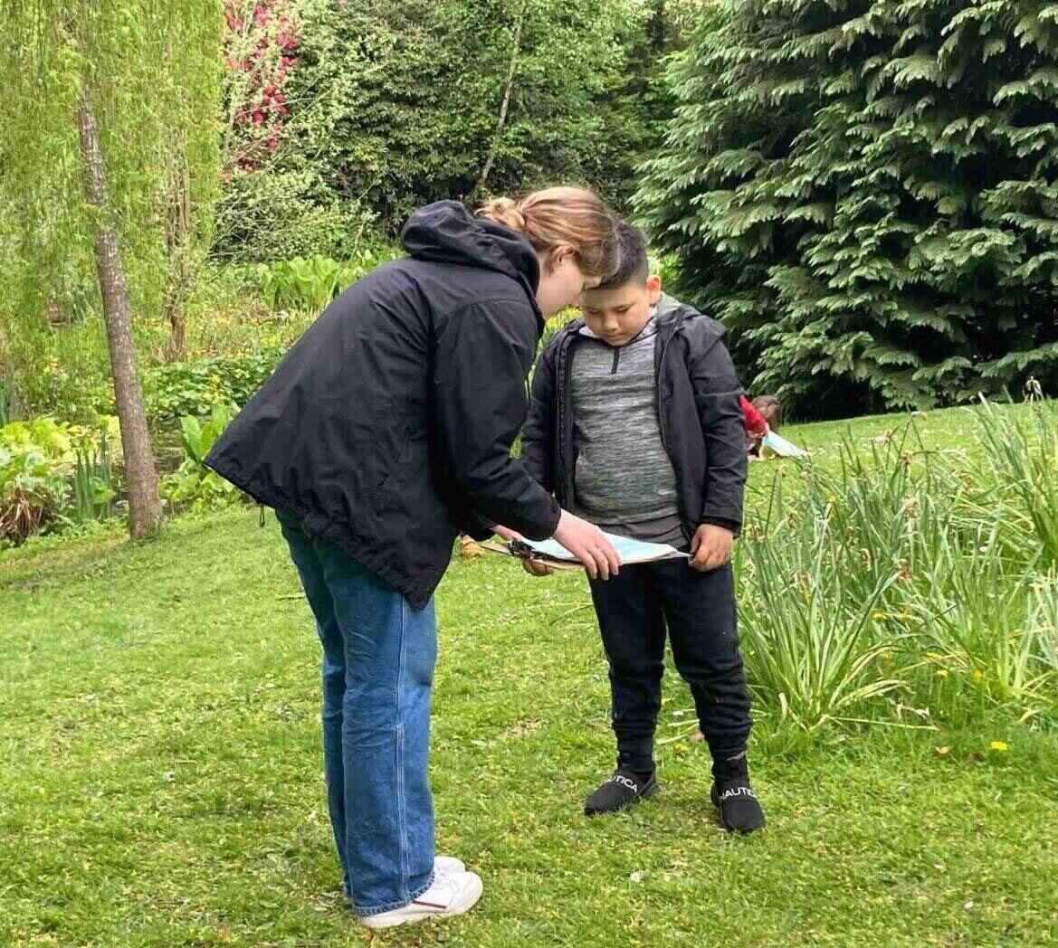 A woman and young boy standing together on grass with trees in the background. They are looking at paper on a clipboard together. 