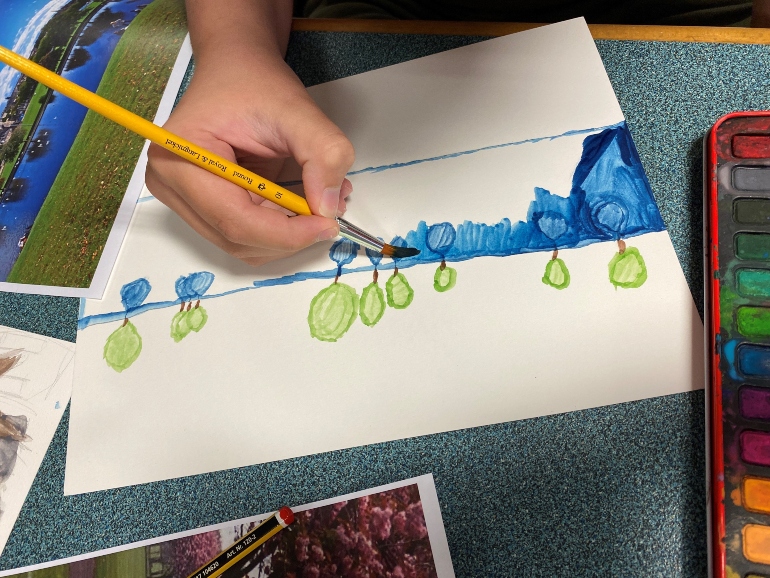 Photograph of child painting blues and greens on a white piece of paper. A colourful paint palette is next to the young artist 