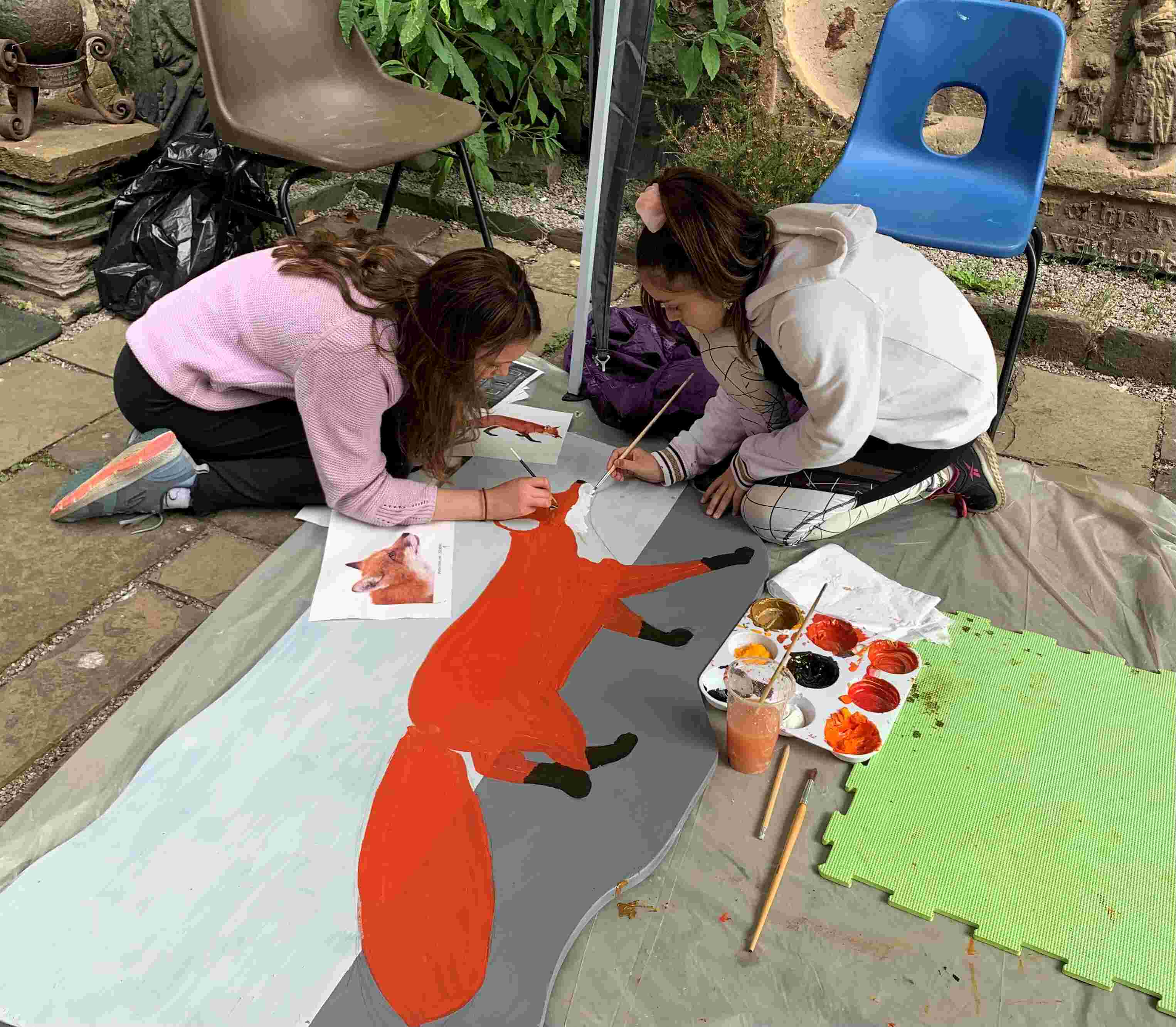 Two girls kneeling on the ground painting a fox on to a wooden board.