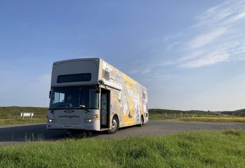 The iconic Travelling Gallery bus parked by the road and surrounded by a green landscape and clear blue sky