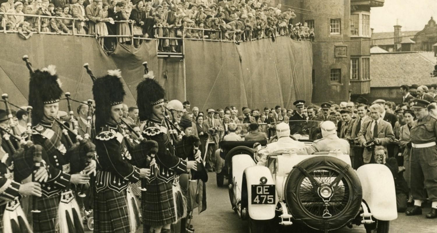 A crowd looking down on two racing cars, each with driver and passenger. Bagpipers stand to the left, more crowds to the right