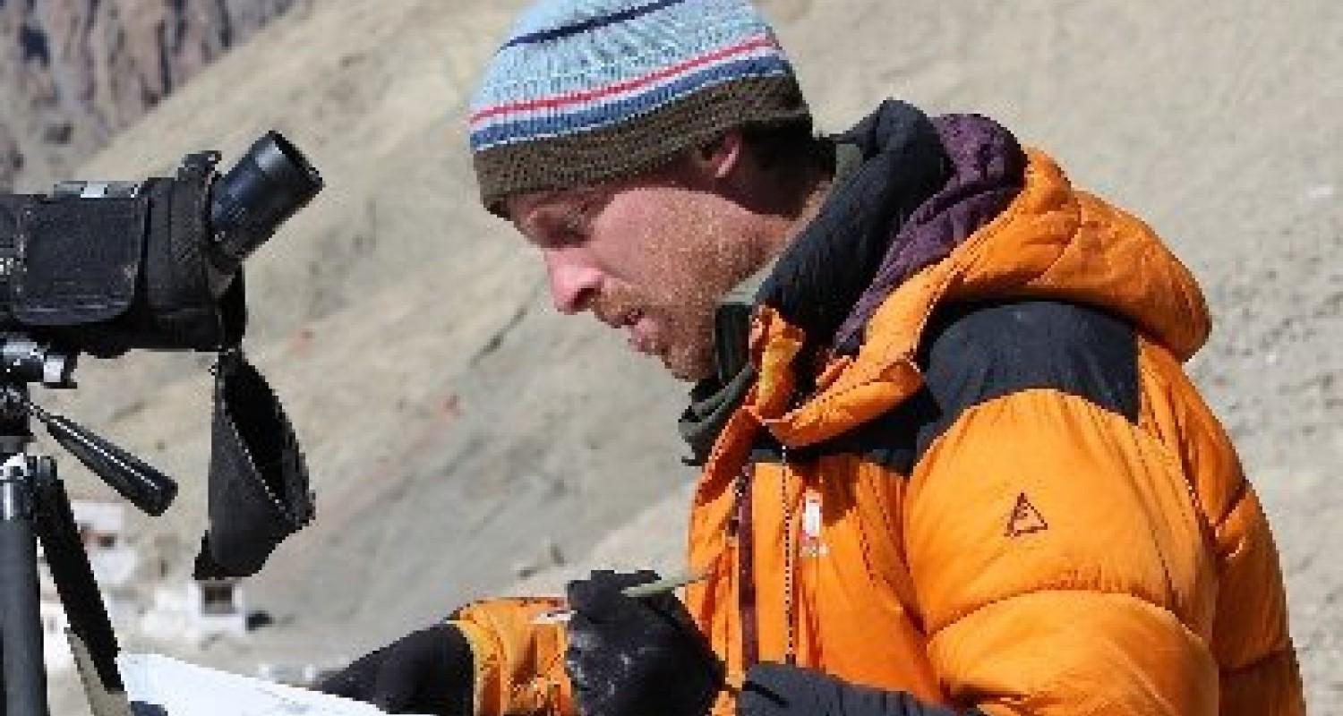 Chris Wallbank in an orange anorak, wearing a stripy beanie, making notes in front of his camera