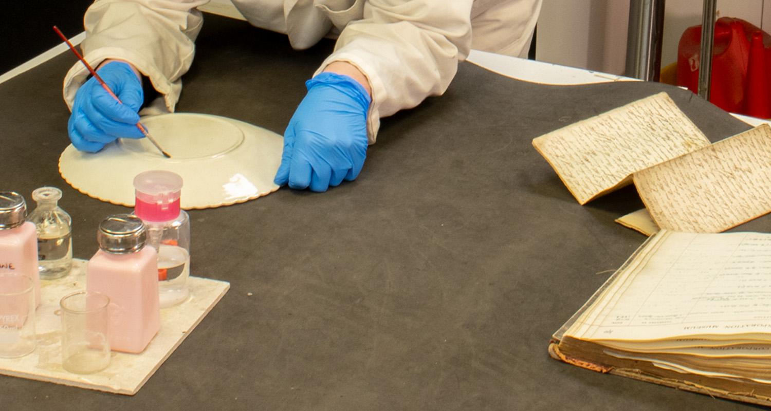 A pair of gloved hands handling a plate at a table with pots of brushes and old books and documents