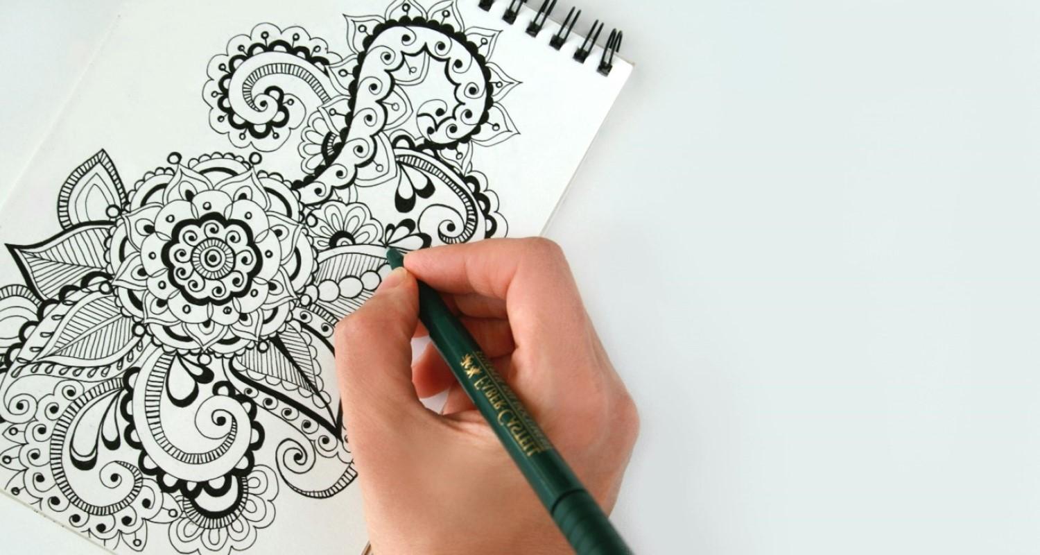 A photo of a hand with a green pen doodling a black and white design of a flower with petals and tentacles  
