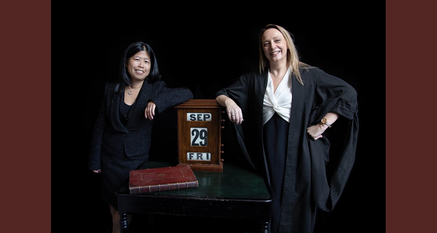 Jennifer Ang and Lynn McMahon in a black suit and black gown respectively leaning on a large wooden calendar which depicts Friday 29 September. A red leather-bound book is in front of it on the dark wood table 