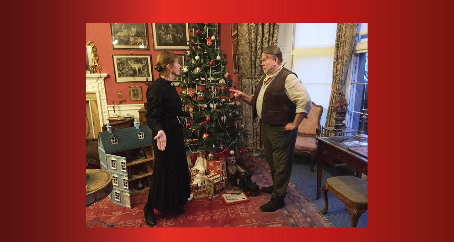 A scene from the tour, with a couple standing talking in a room at Lauriston in front of the Christmas tree, a dollshouse and a selection of gifts