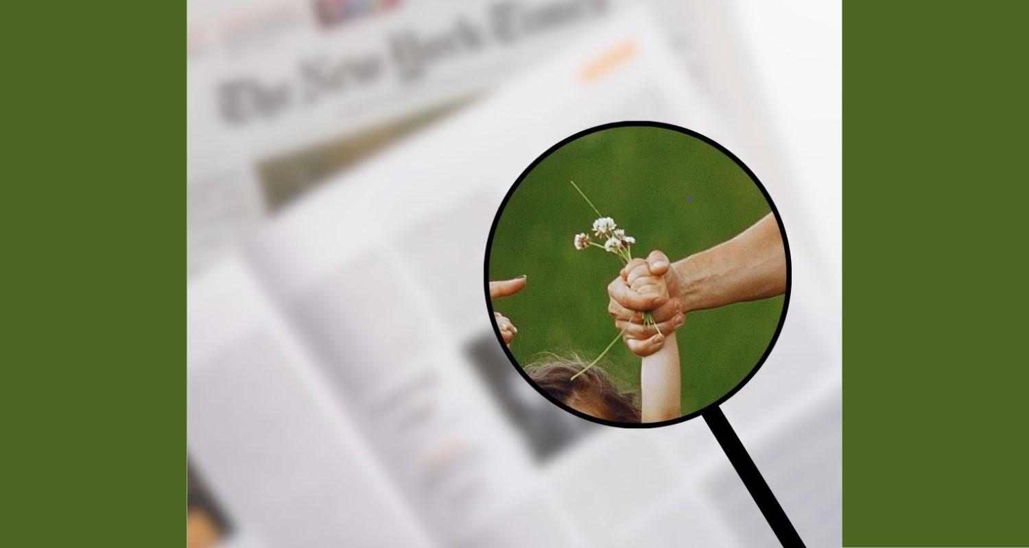 A magnified image of a child's hand holding a flower, held by an adult's hand. Newspapers beneath are out of focus.