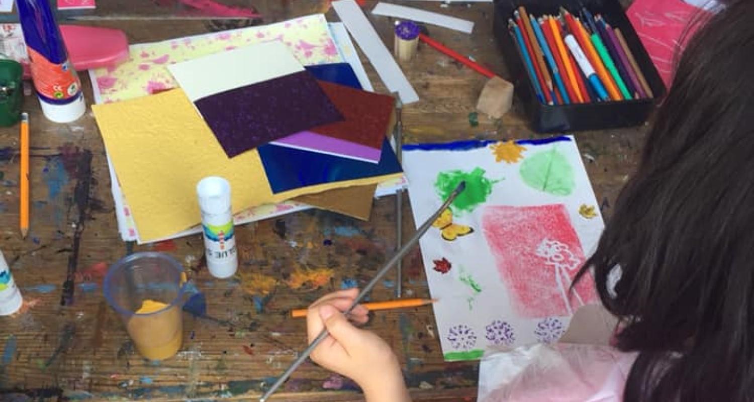 A child painting a picture with leaves, a butterfly and flowers. The desk is covered in art materials.
