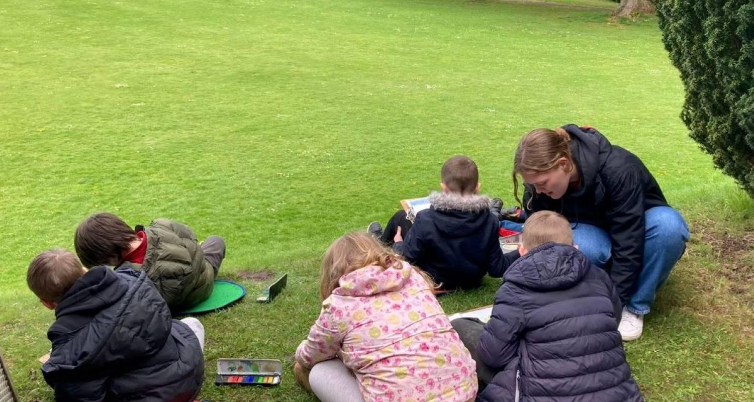 A volunteer helping a group of children sitting on grass with paper and paints