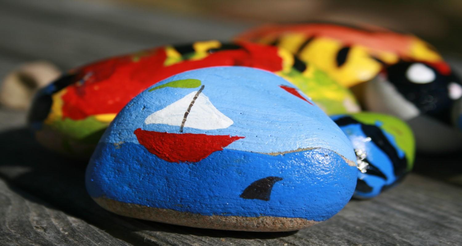 A painted rock showing a red yacht with a white sail and green flag and a shark in the water nearby