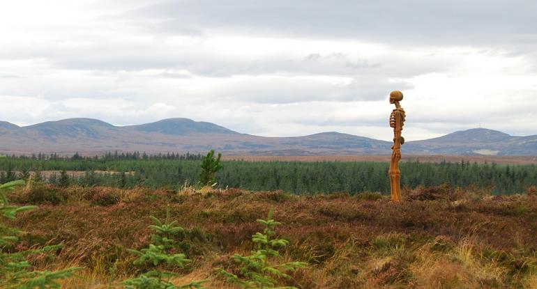 A skeleton stands on the edge of Borgie Forest in Sutherland. There are mountains and a grey, cloudy sky in the background