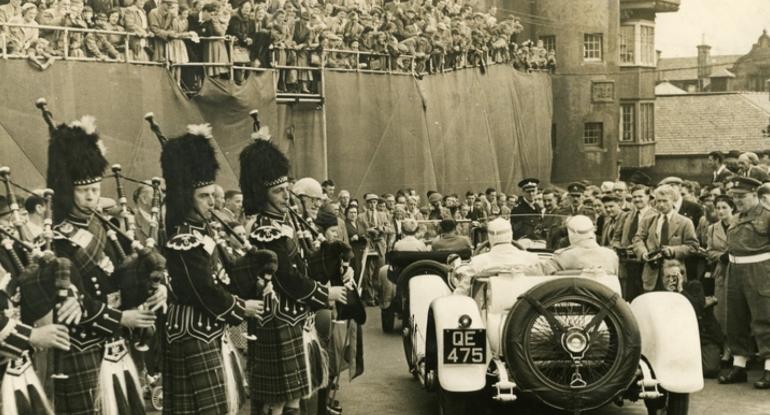 1930s racing cars A crowd looking down on two racing cars, each with driver and passenger. Bagpipers stand to the left, more crowds to the right