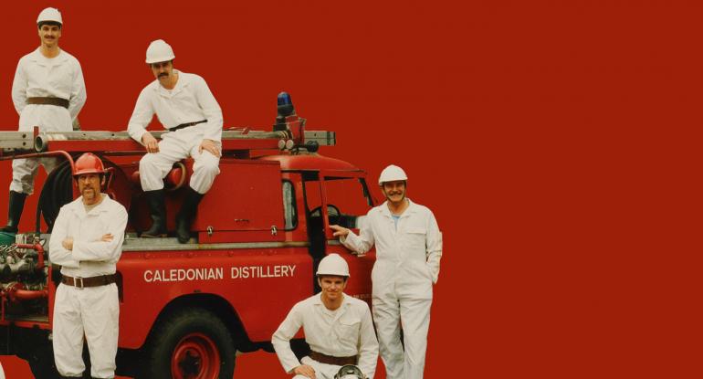 Detail of members of the Caledonian Distillery Fire Brigade against a plain red background
