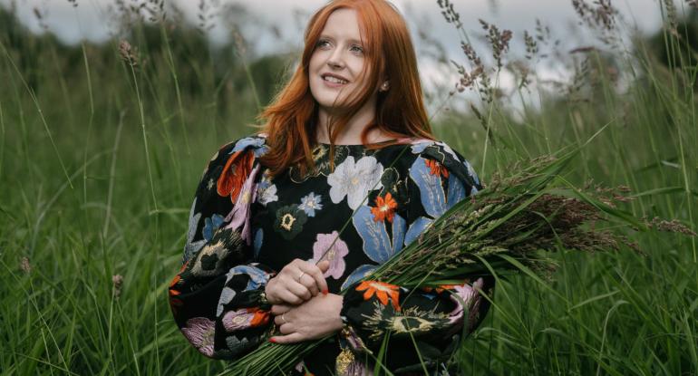 Ceitlin Lilidh sits in a green meadow wearing a black dress with large bold and colourful flowers