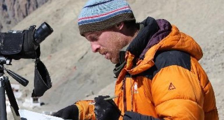 Chris Wallbank in an orange anorak, wearing a stripy beanie, making notes in front of his camera