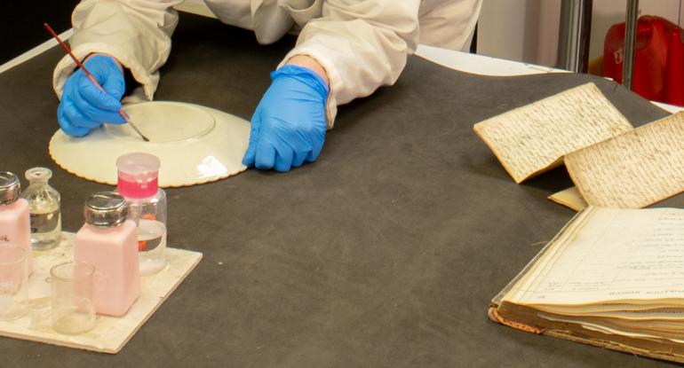 A pair of gloved hands handling a plate at a table with pots of brushes and old books and documents