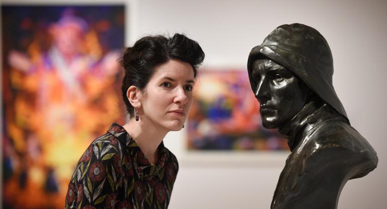 Curator in a gallery at the City Art Centre in front of a Sculpture with paintings behind her. 