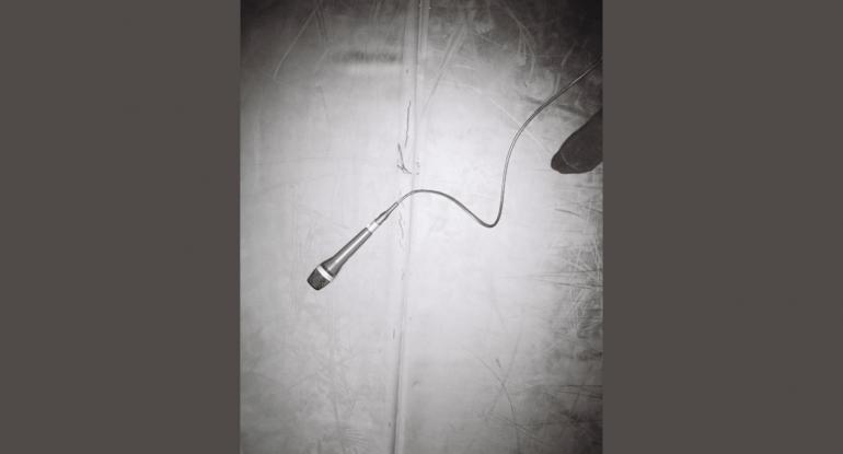 A black and white photograph of a microphone on a cord lying on the floor; a socked foot is visible top right