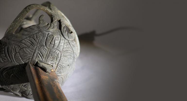 A close up of a sword hilt and blade of the type known as a mortuary, with an engraved stylised head.