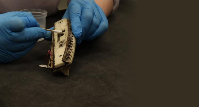 A close up image of a hand in blue gloves cleaning a small model boat with a cotton bud