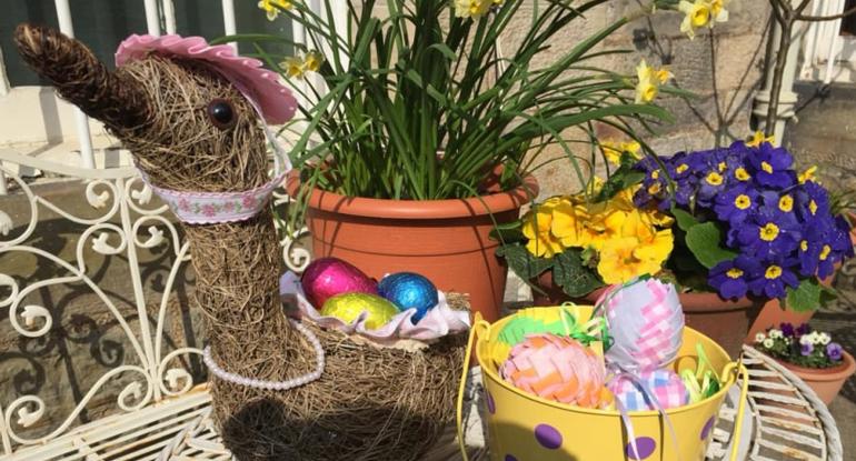 A wicker duck with easter eggs in a yellow bucket and flowerpots in the background