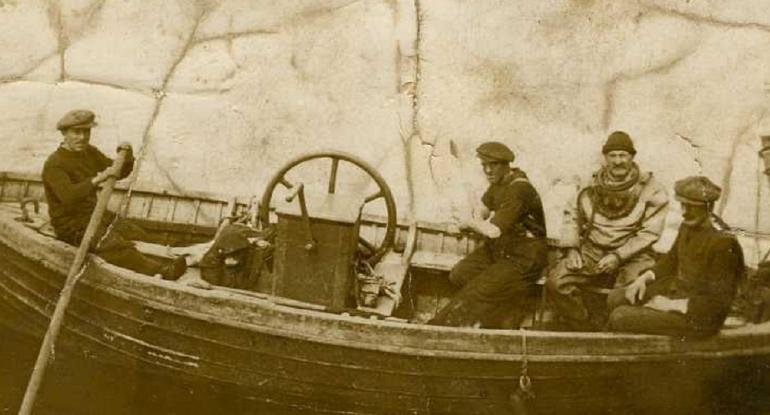 Sepia photograph of four men in a boat, one in a full diving suit.