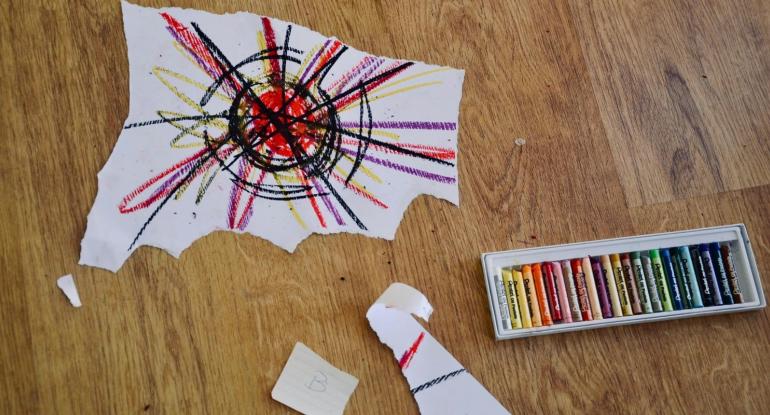 Ripped, crayon-ed pieces of paper and a box of coloured crayons on a desk