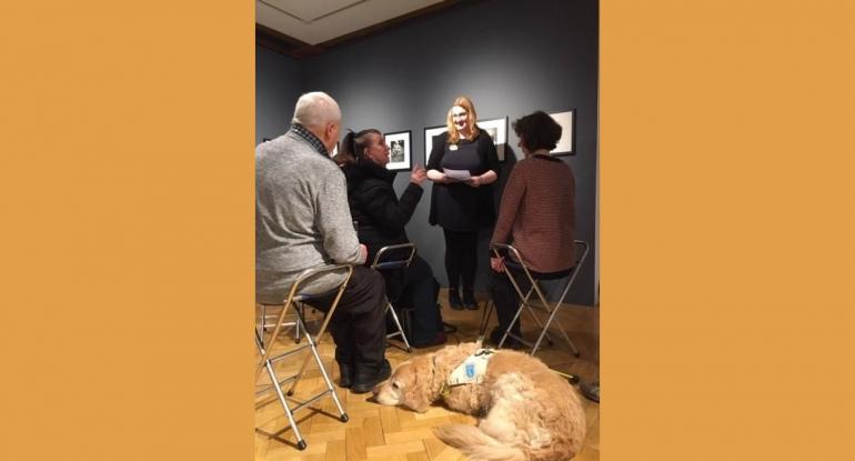 Described tour Tessa standing in the City Art Centre in front of black and white photos, with visitors to a Described tour - and a snoozing Golden Retriever