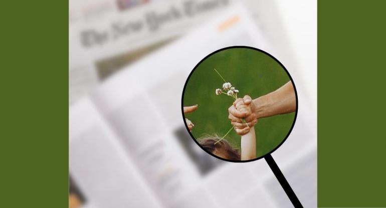 A magnified image of a child's hand holding a flower, held by an adult's hand. Newspapers beneath are out of focus.