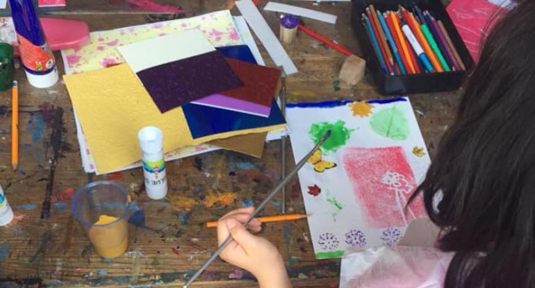 A child painting a picture with leaves, a butterfly and flowers. The desk is covered in art materials.