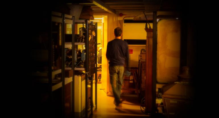 A man seen from the back walking through a dark museum store towards an open door into a lit room
