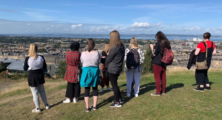 A group of young people stand on a hill in Edinburgh and face Scotland's capital below