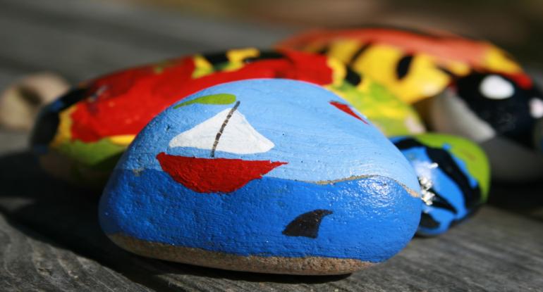 A painted rock showing a red yacht with a white sail and green flag and a shark in the water nearby