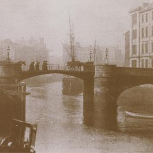 A dark image of a drawbridge with people walking over it across a canal.