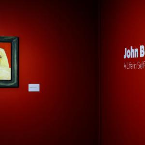 Framed artwork on a red wall and the name of the art exhibition on the opposite wall - John Bellany: A Life in Self-Portraiture