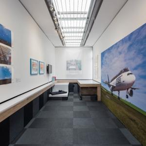 Gallery interior. 5 photographs are pinned to a wall and opposite is a large photograph of a plane on a runway.