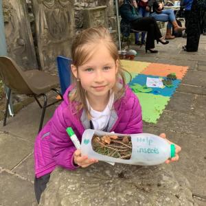 A girl in a purple coat holding a plastic milk bottle that has been turned into a bug hotel with 'save the insects' written on it