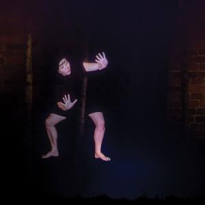 Woman as ghostly figure posing in a dark setting in front of a brick wall.