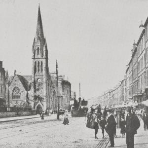 A busy Leith Walk street scene with a motorised tram in the distance and a church in the background.
