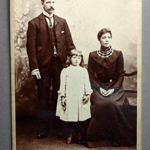 A cabinet card by Pettigrew & Amos showing a man with moustache, woman in a high necked, long sleeve black dress and small girl in a white dress.